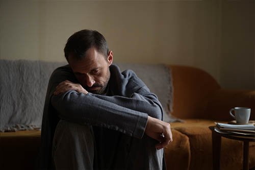Мужчина сидит на полу грустно смотрит вниз, обнимает колени